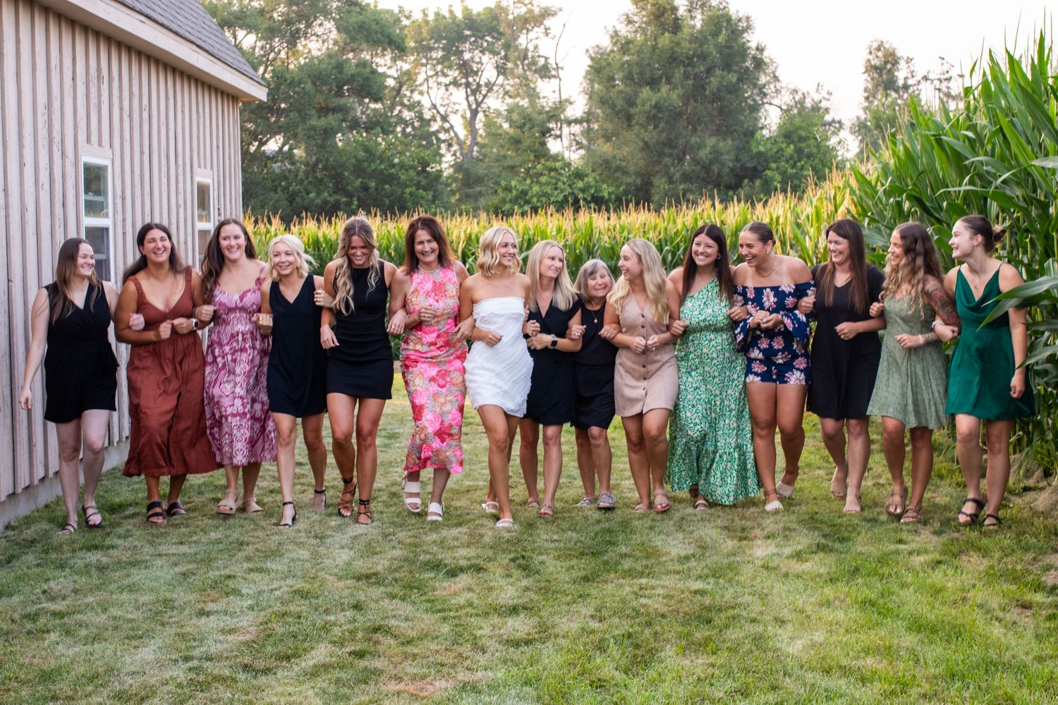 A group of women in colorful dresses laugh and walk together outdoors near a barn and cornfield.