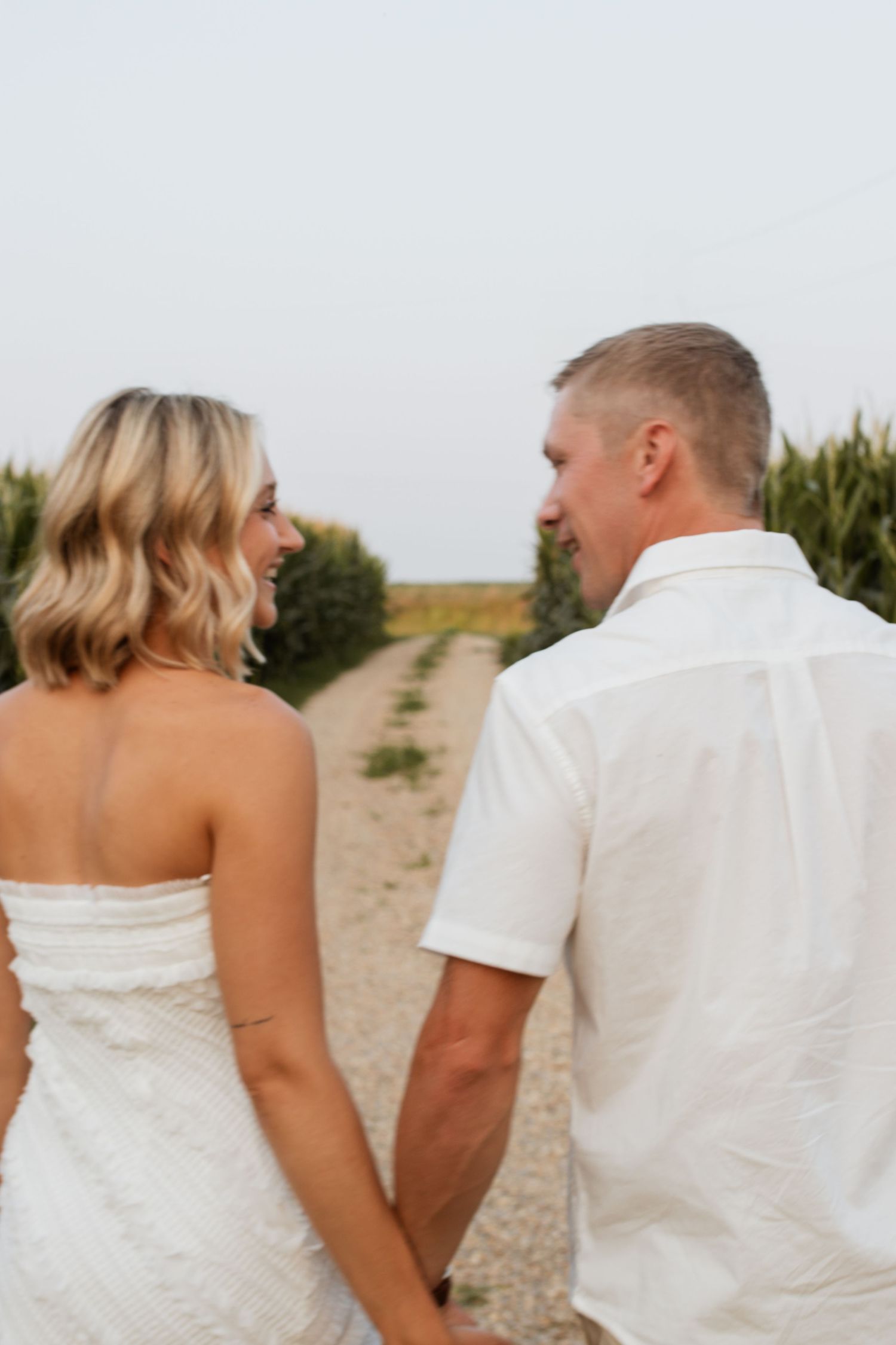 Couple in white attire holding hands walking down a rural path, woman looking back smiling.