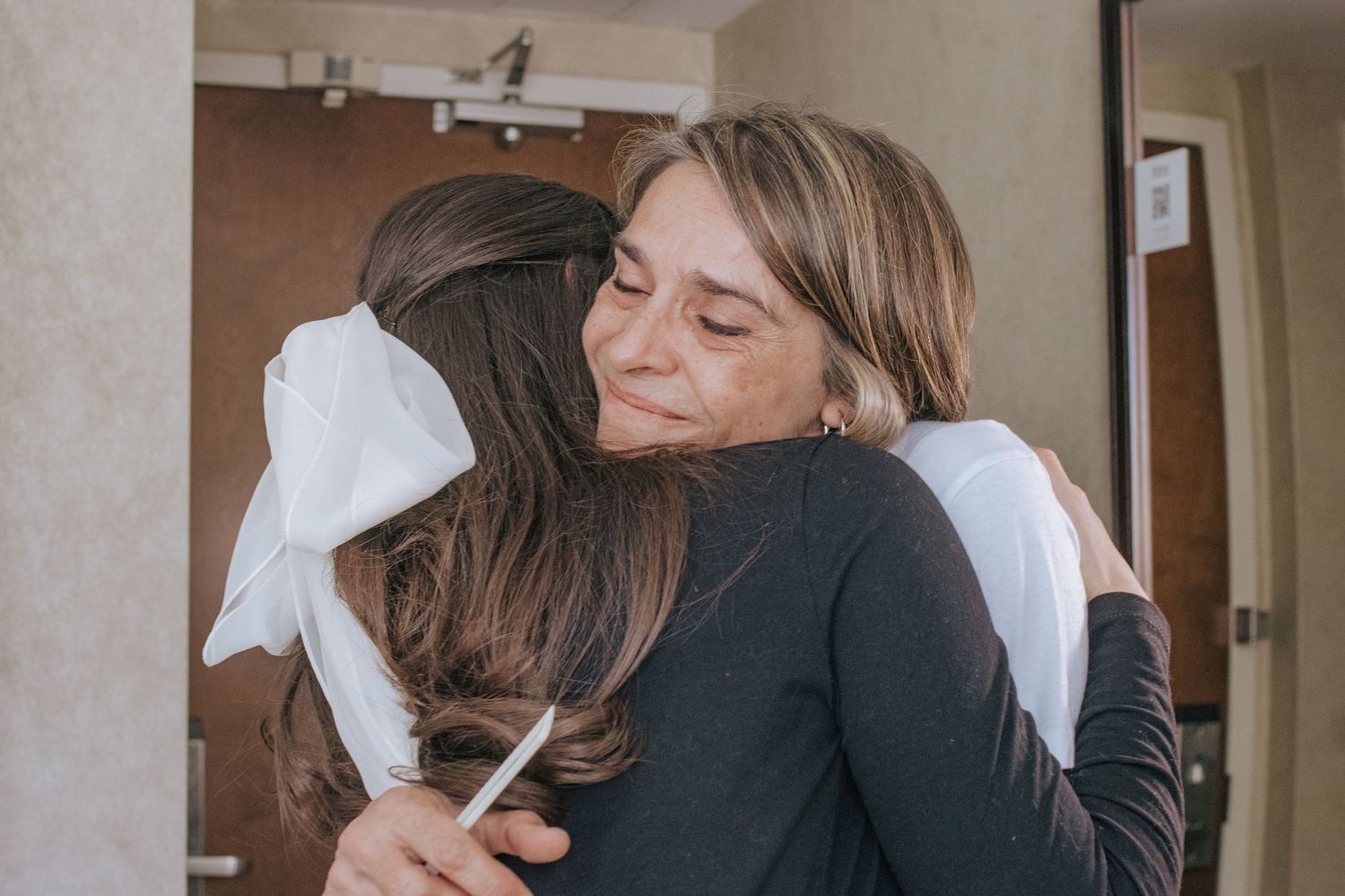 Two women sharing a warm emotional hug, one wearing a large white bow in her hair.
