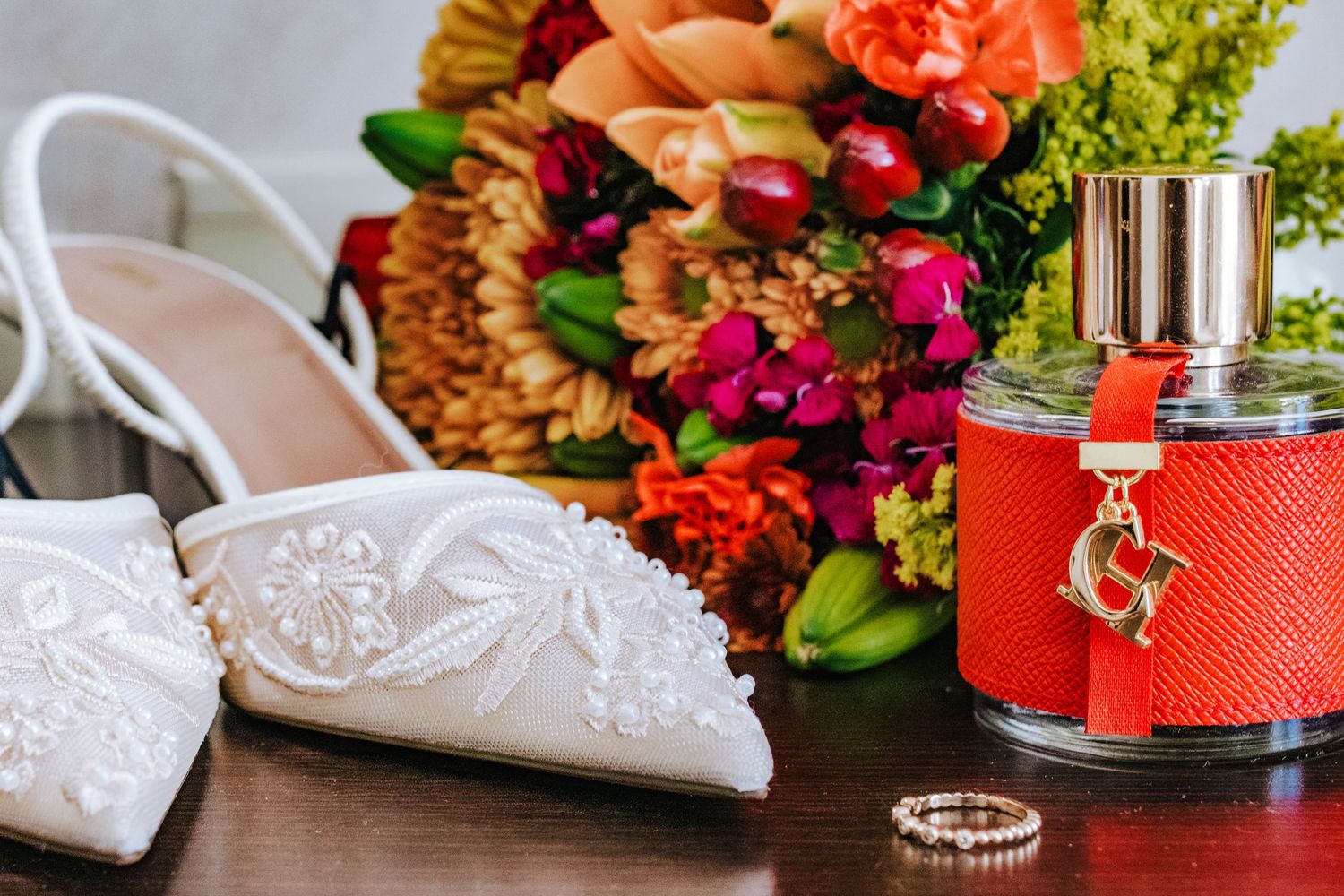 White embroidered bridal heels, colorful wedding bouquet, red perfume bottle, and diamond rings arranged on a wooden surface.