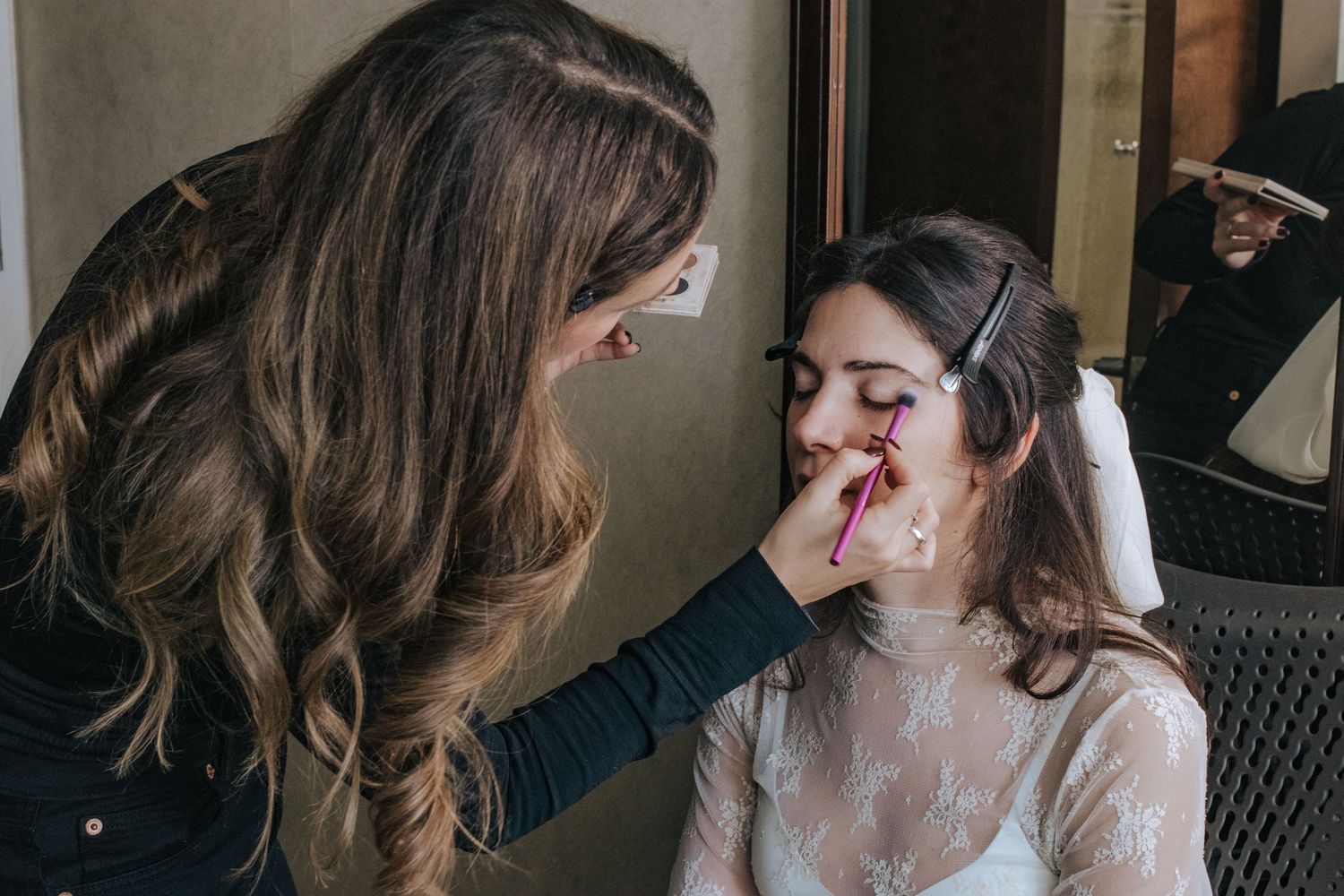 A makeup artist applies lipstick to a bride in a white lace dress, preparing her for her wedding day.