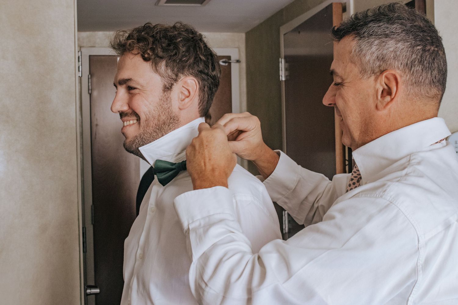 A man helps a groom tie a green bow tie while preparing for a wedding ceremony indoors.