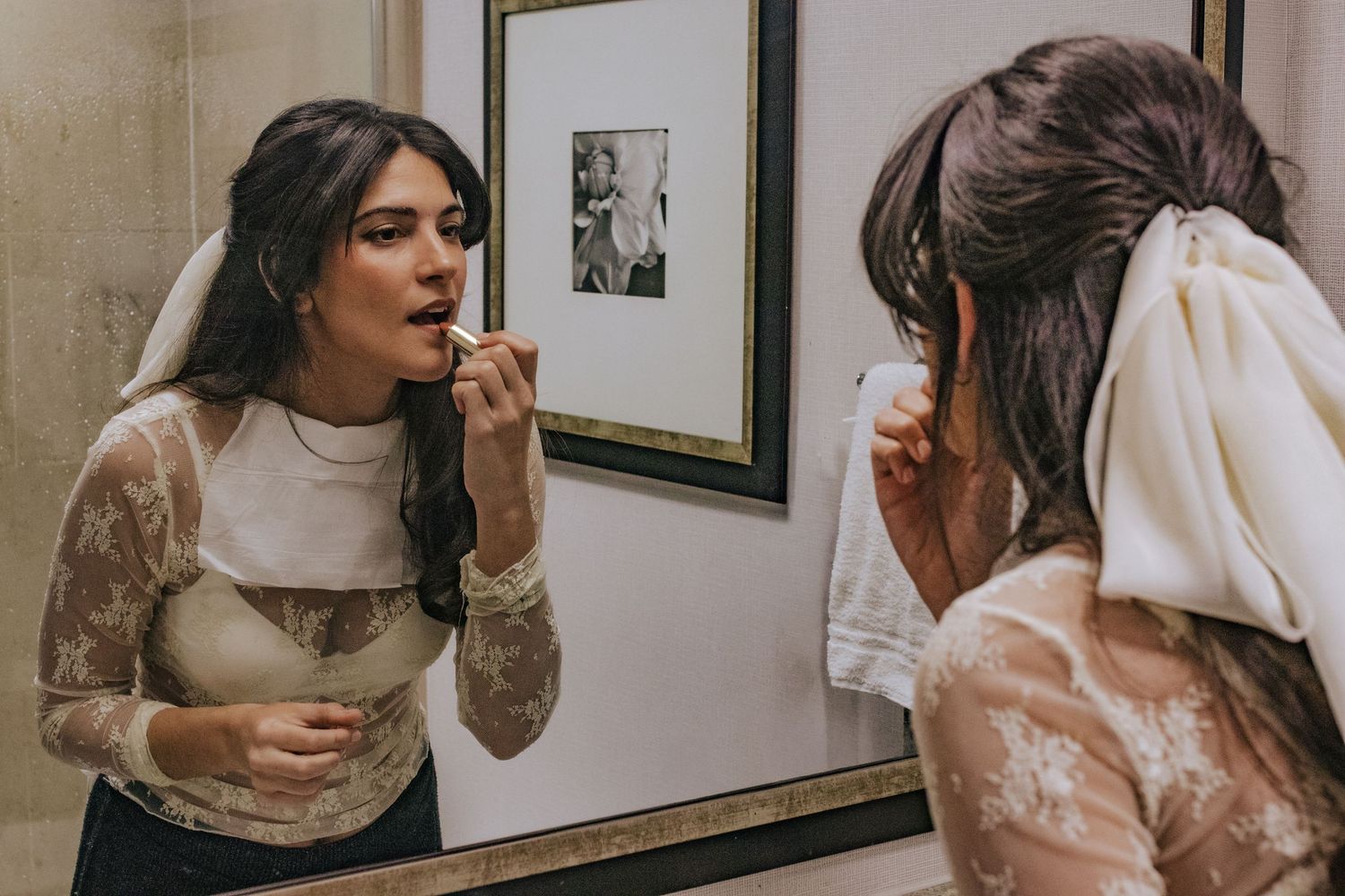 Bride in lace wedding dress applying lipstick while looking in bathroom mirror before her wedding ceremony.
