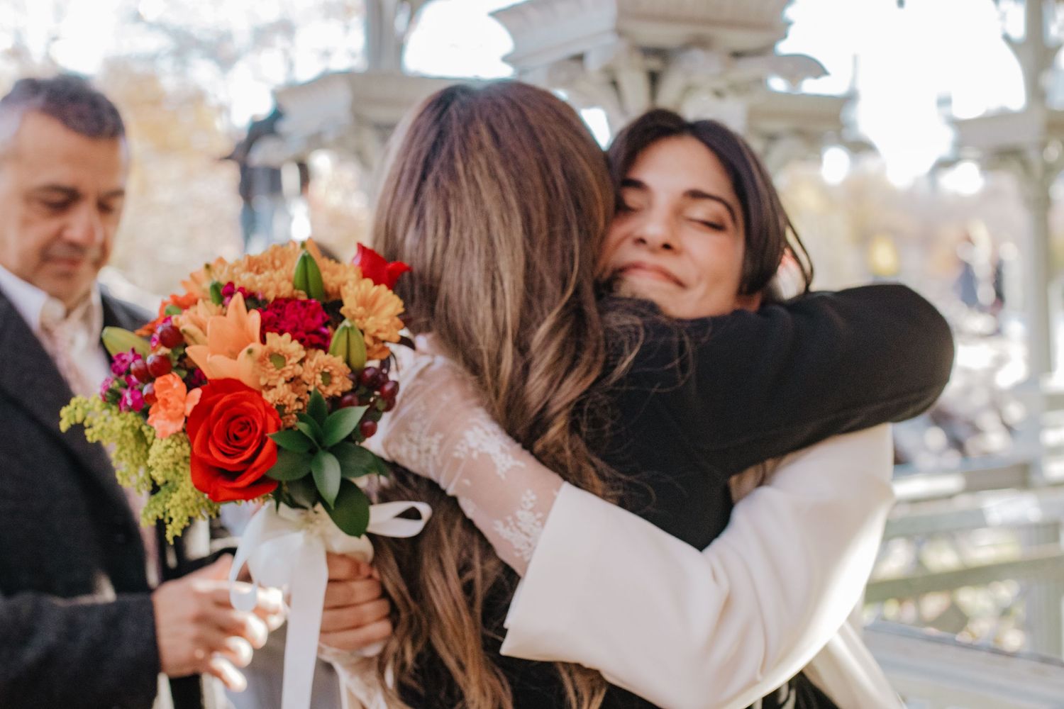 Two women share a warm embrace at an outdoor wedding celebration, one holding a vibrant bouquet of red and orange flowers.