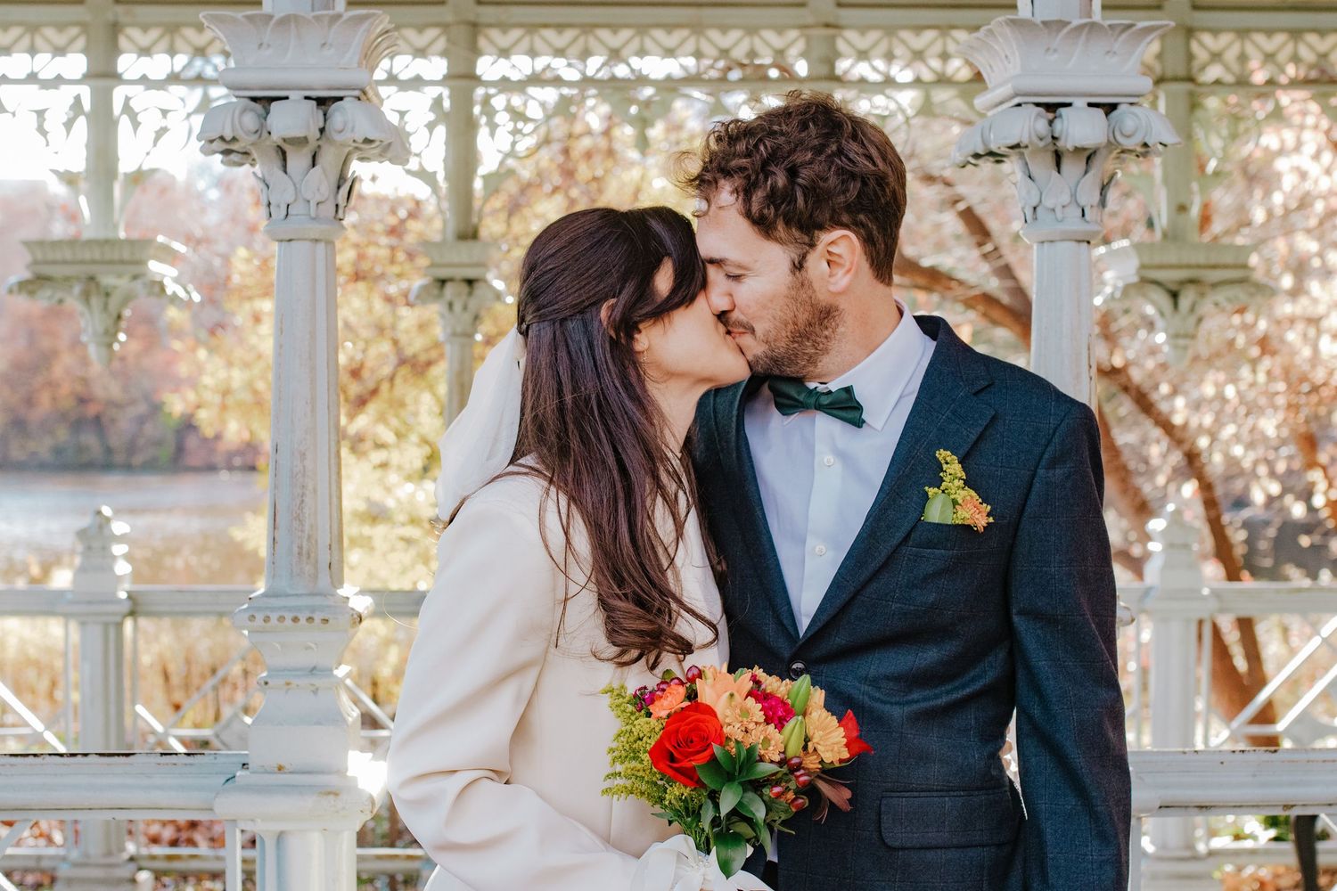 Bride and groom sharing a kiss at Central Park gazebo, bride holding vibrant bouquet, groom in navy suit with bow tie.
