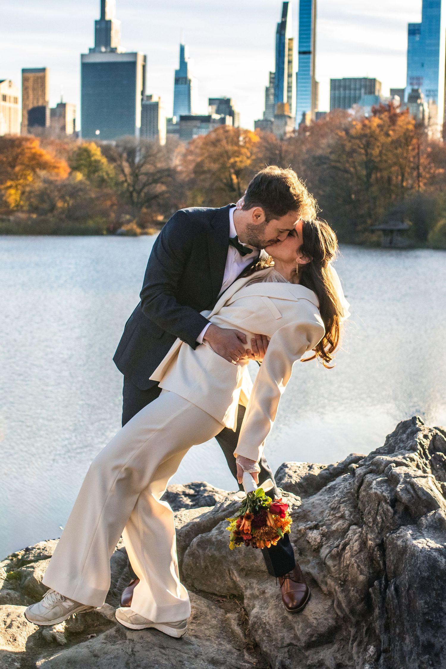 Couple sharing a romantic dip kiss in wedding attire with Central Park lake and NYC skyline in autumn background.