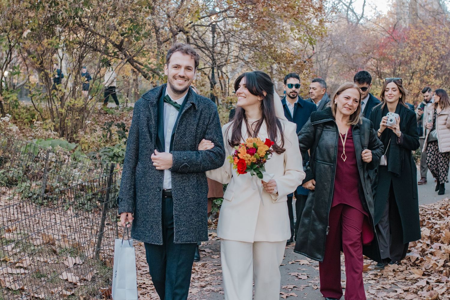 Couple walking in autumn park, woman holding colorful bouquet, guests following behind on tree-lined path.