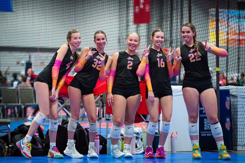 Five young female volleyball players in black uniforms smiling together on an indoor court during a tournament.