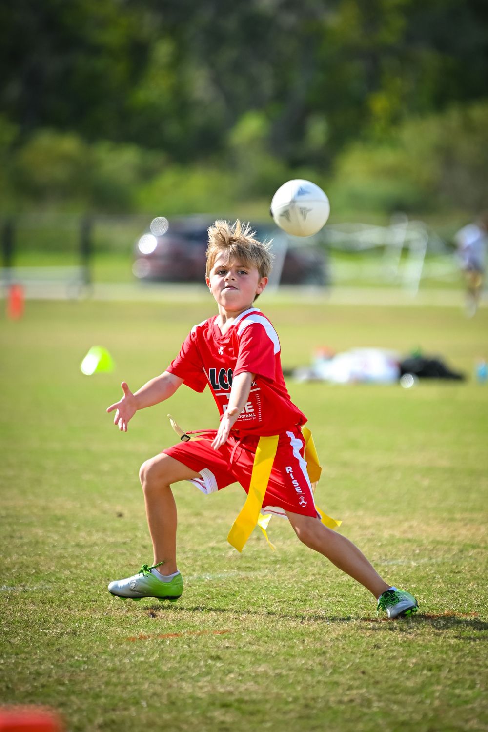 Young boy in red soccer uniform heading a ball during a youth flag football game on a sunny green field.