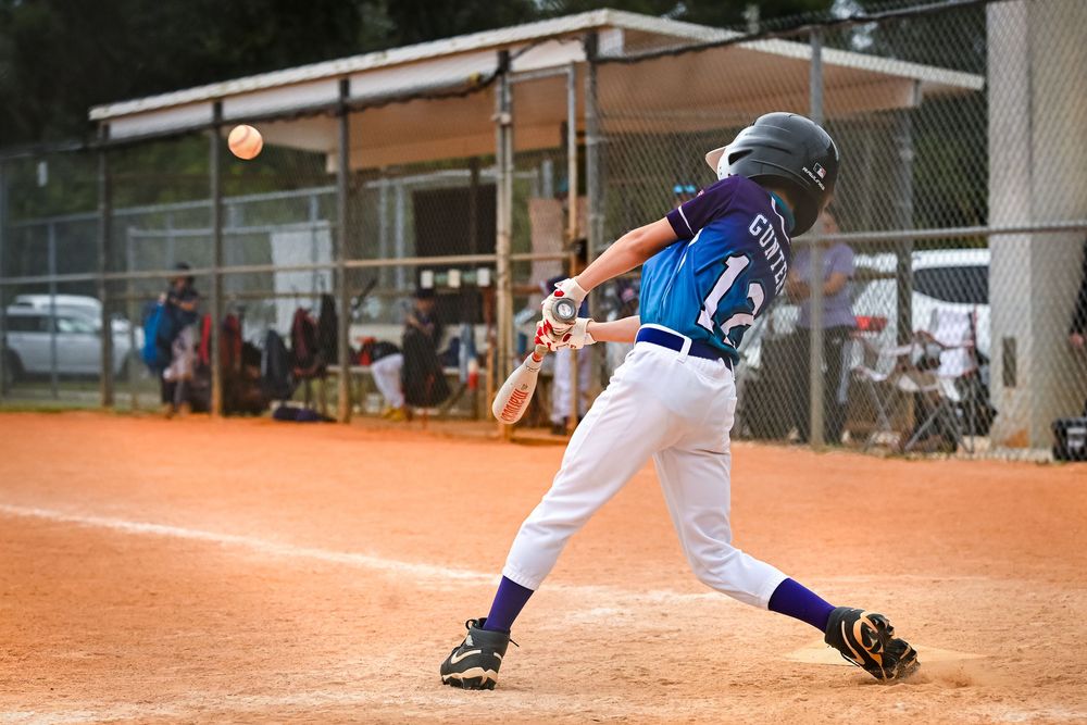 Young baseball player in white uniform swinging bat at incoming pitch during a youth league game on a dirt field.