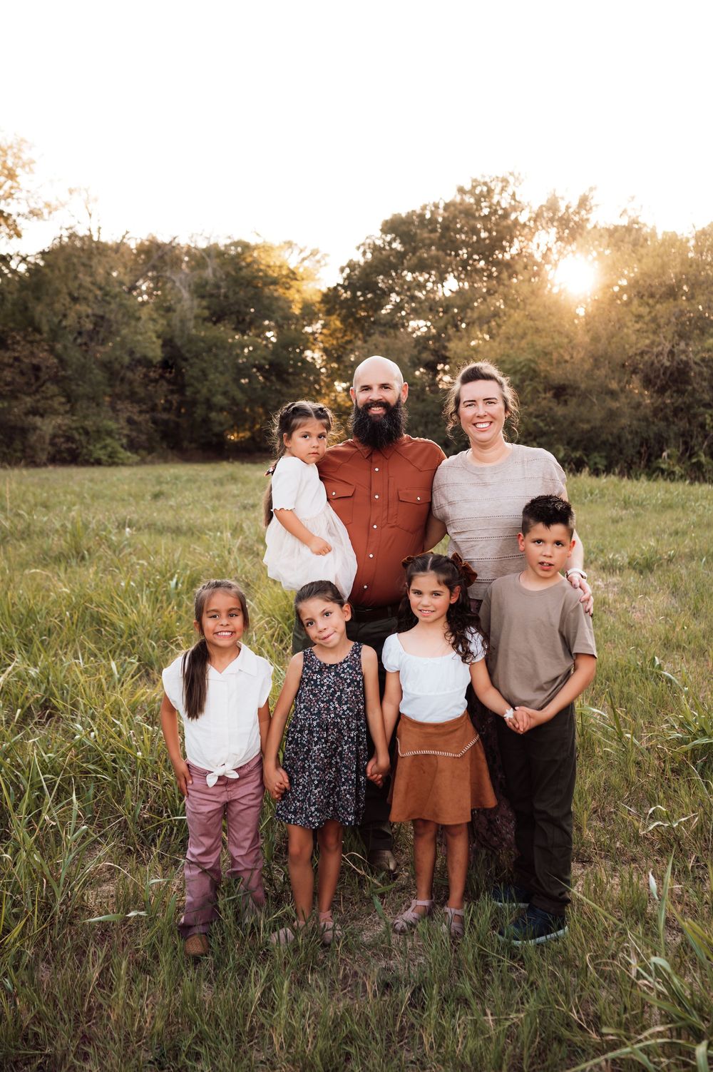 Family of seven poses in a sunlit green meadow at golden hour, with parents and five children smiling outdoors.
