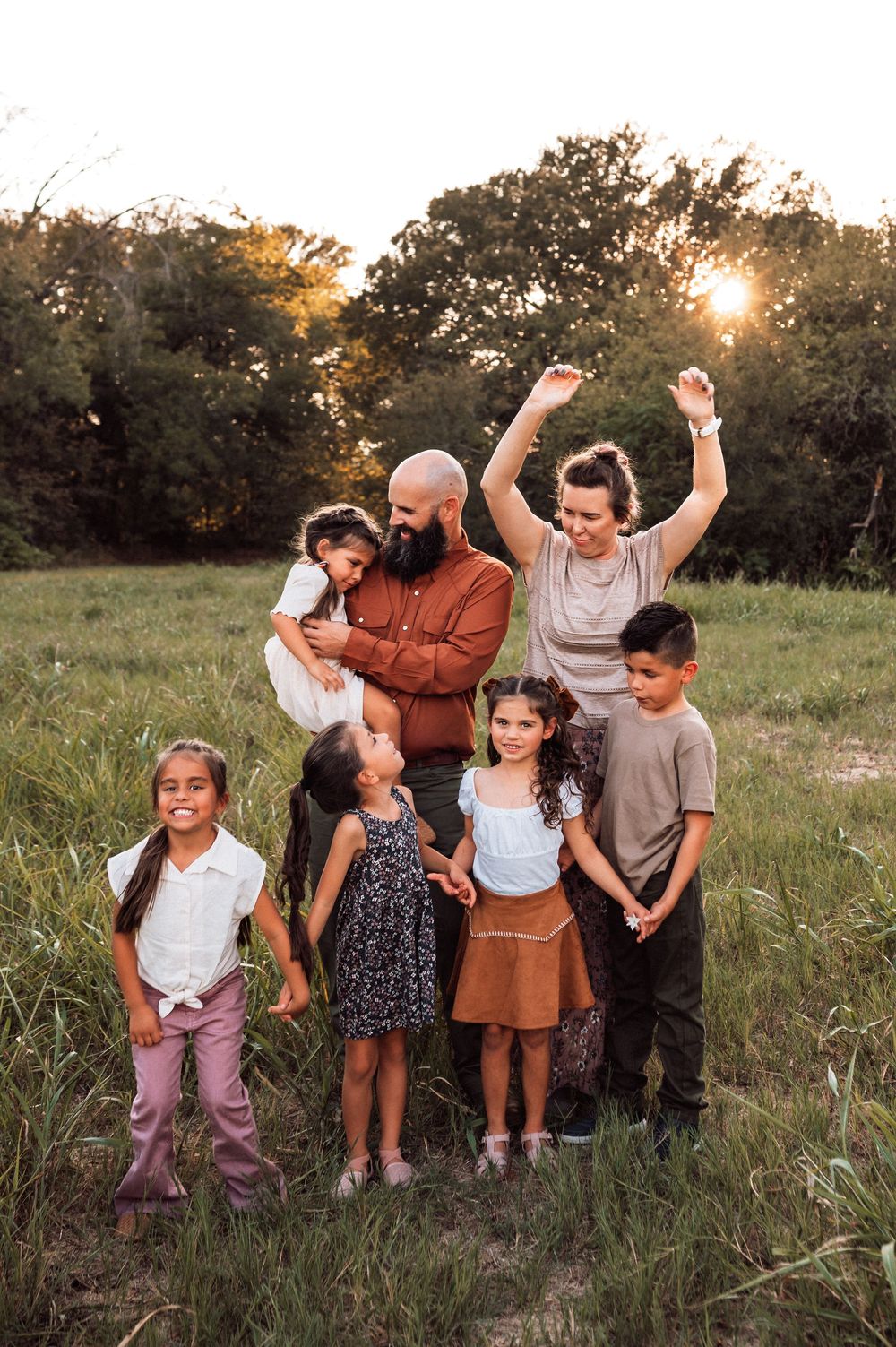 A joyful family of seven poses in a sunlit field at golden hour, with parents raising hands and children smiling.