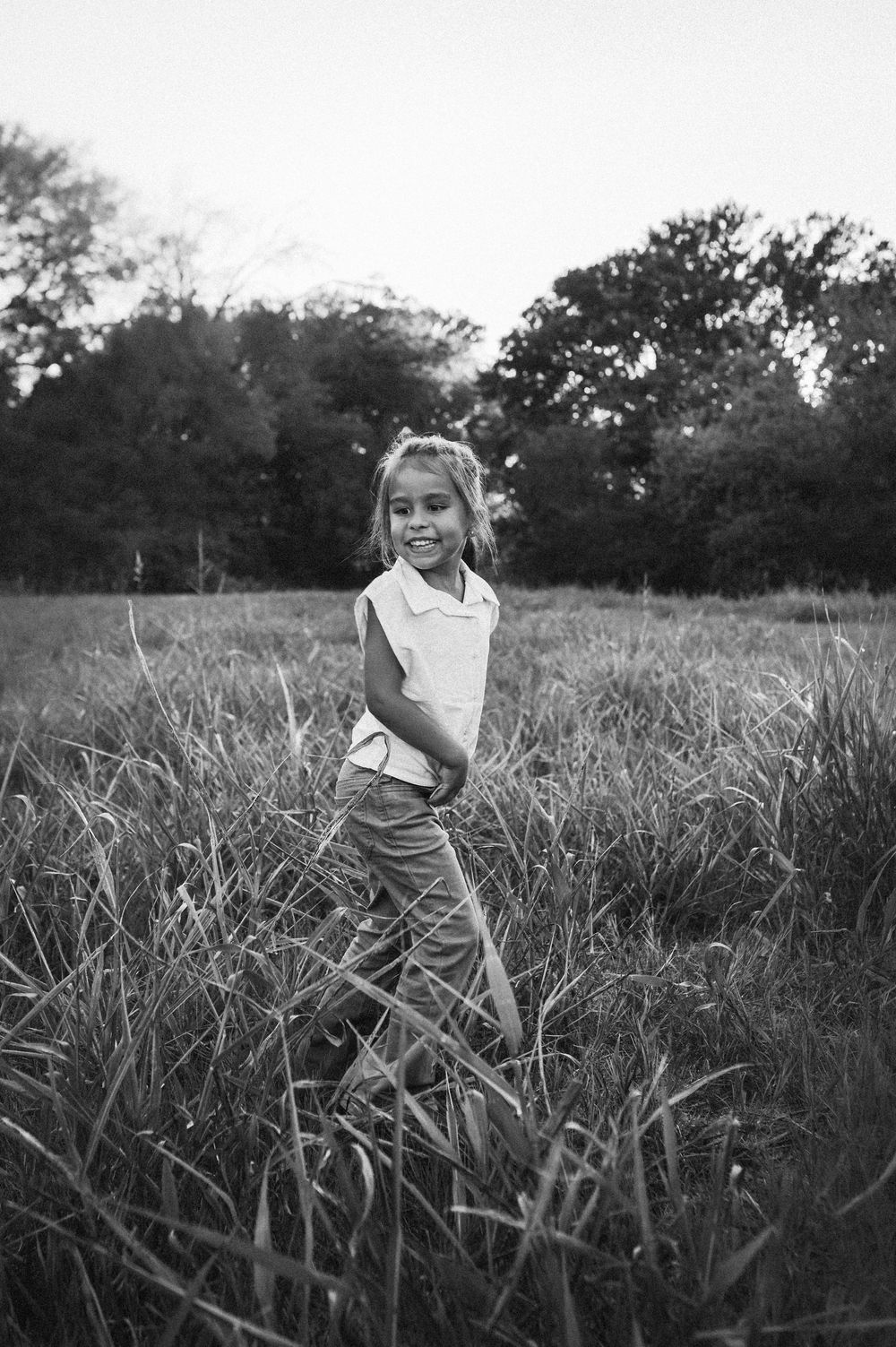 Black and white photo of a smiling young girl standing in tall grass field with trees in background.