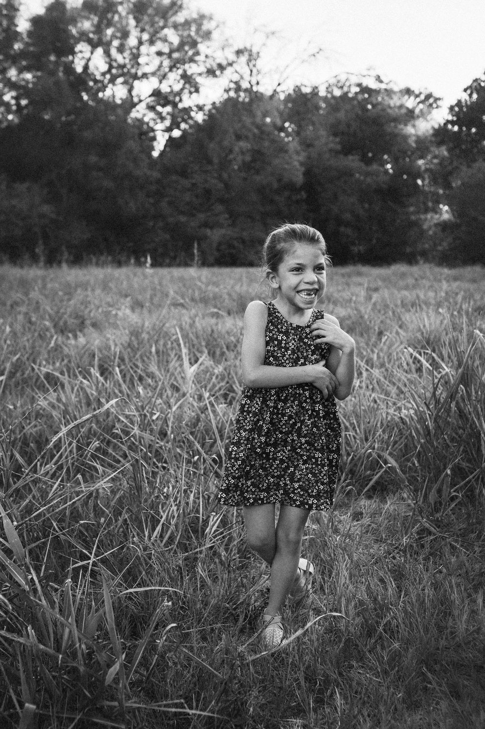 A joyful young girl in a floral dress laughs while running through a grassy field with trees in the background.