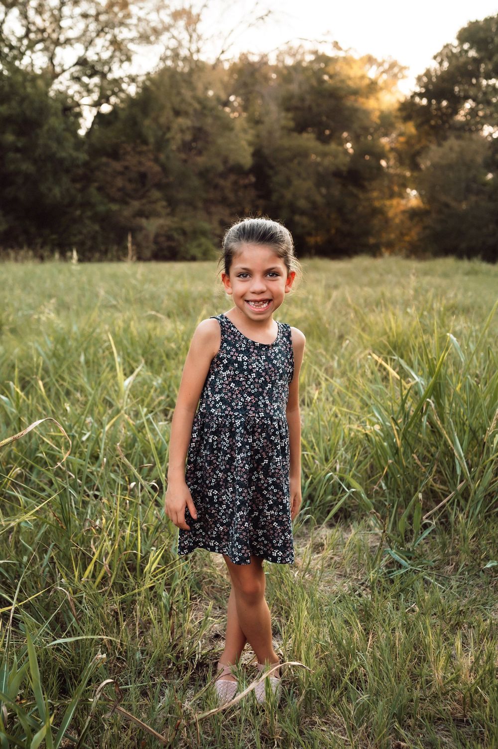 A smiling young girl in a floral dress stands in a lush green field at golden hour.