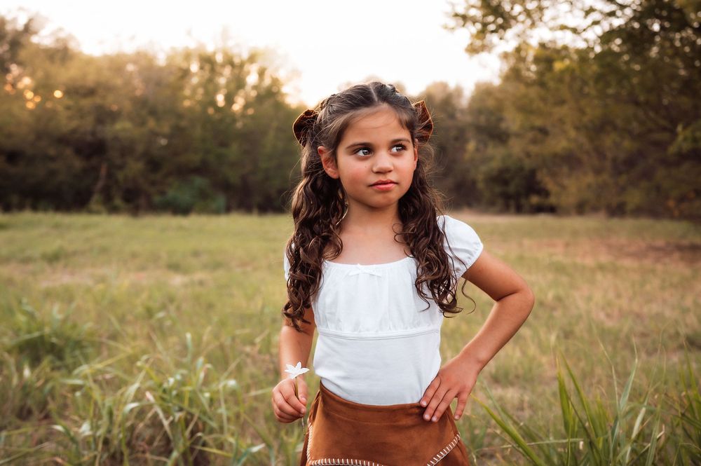 Young girl with curly pigtails in white top and brown skirt posing confidently in a sunlit green meadow.