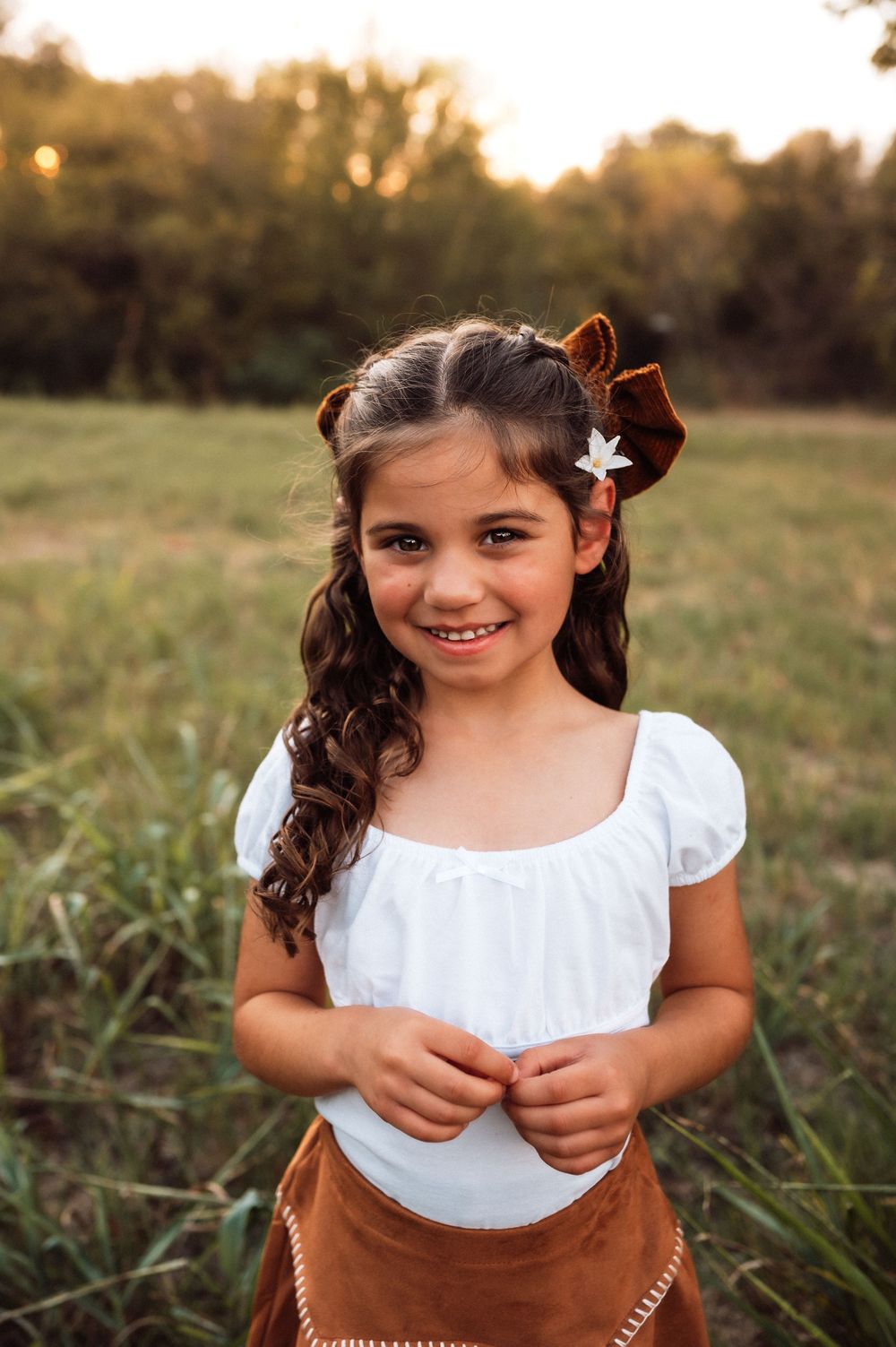 Young girl in white top and brown skirt smiles in a sunny field, wearing brown velvet hair bows.