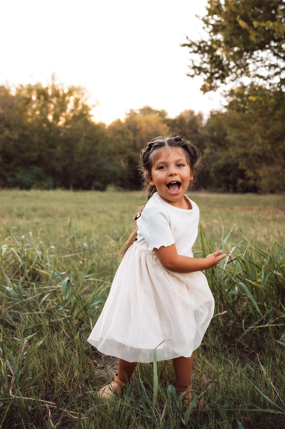 A joyful little girl in a white dress laughs while standing in a green field at golden hour.