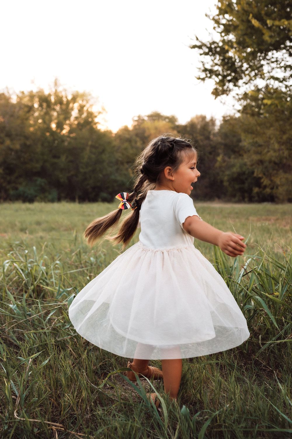 A young girl in a white tulle dress twirls in a sunlit meadow, her pigtail flying as she dances outdoors.