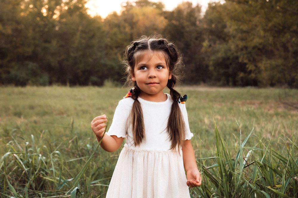 A young girl in a white dress with braided pigtails stands in a green field, making a cute expression.