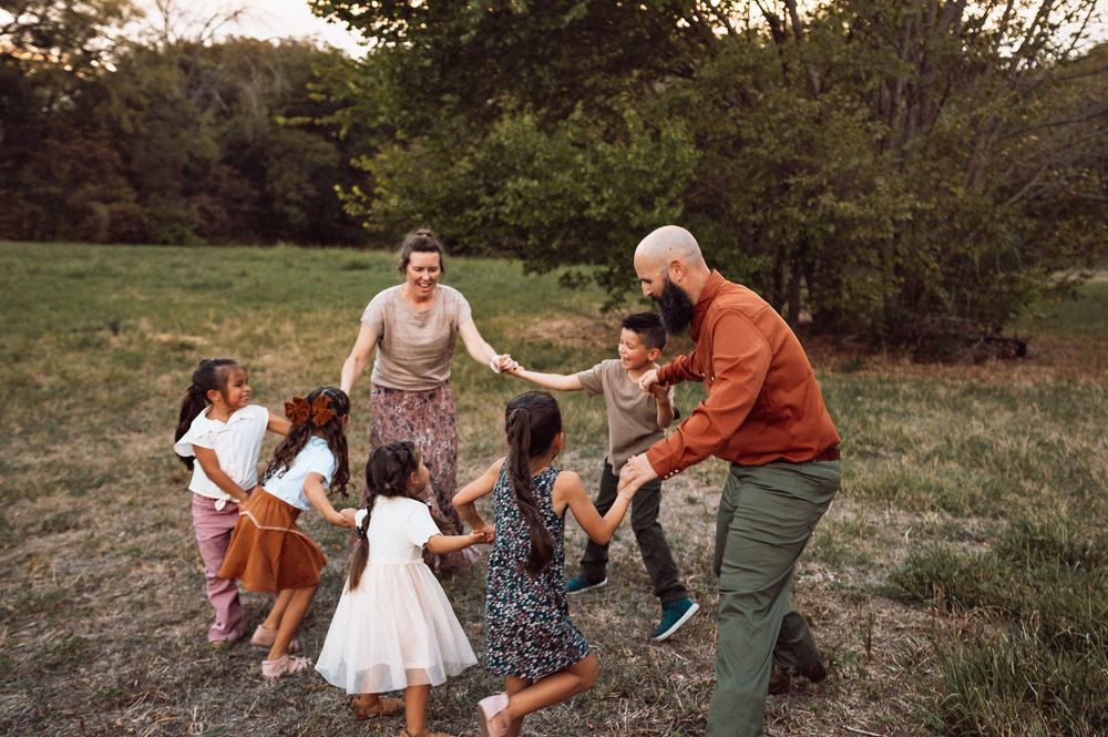 Family with children holding hands in a circle playing outdoors in a green field surrounded by trees.