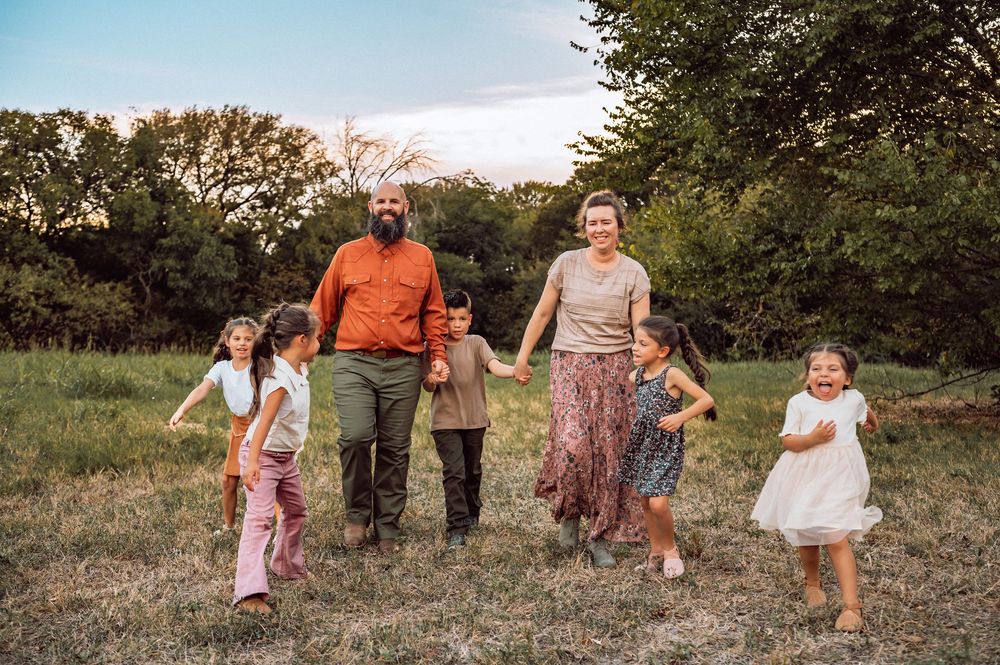 A joyful family with four young children walks through a lush green meadow at sunset, smiling and holding hands.