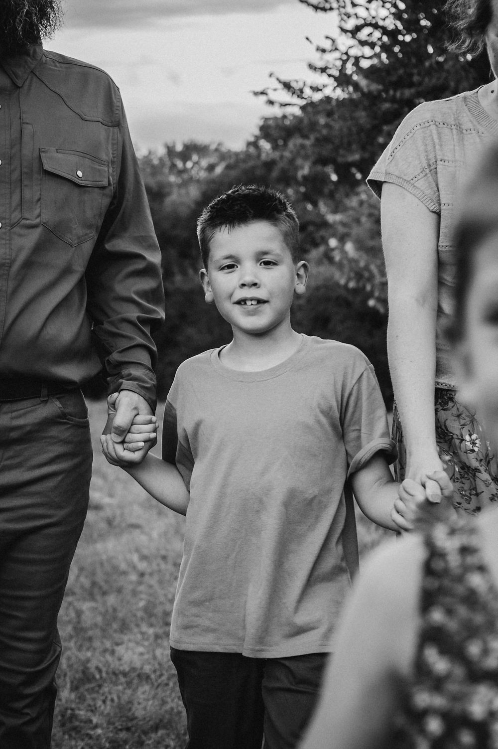Black and white photo of a smiling boy holding hands with parents outdoors among trees.