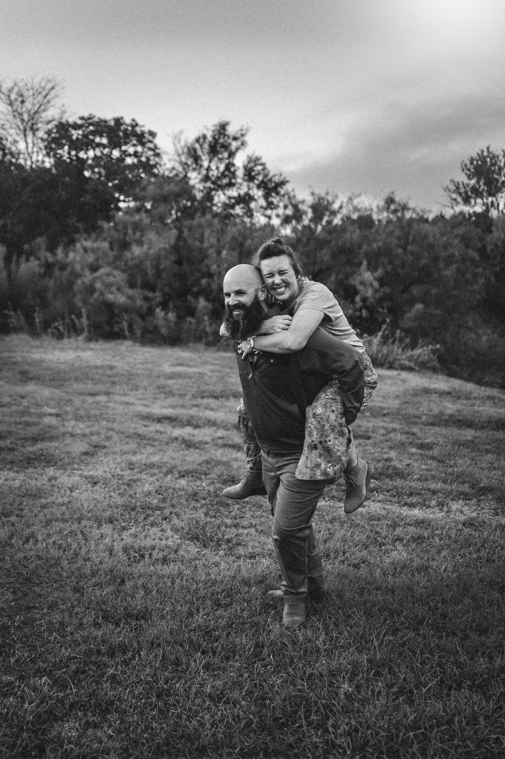 A man gives a woman a piggyback ride in a grassy field, both smiling, in a black and white outdoor photo.
