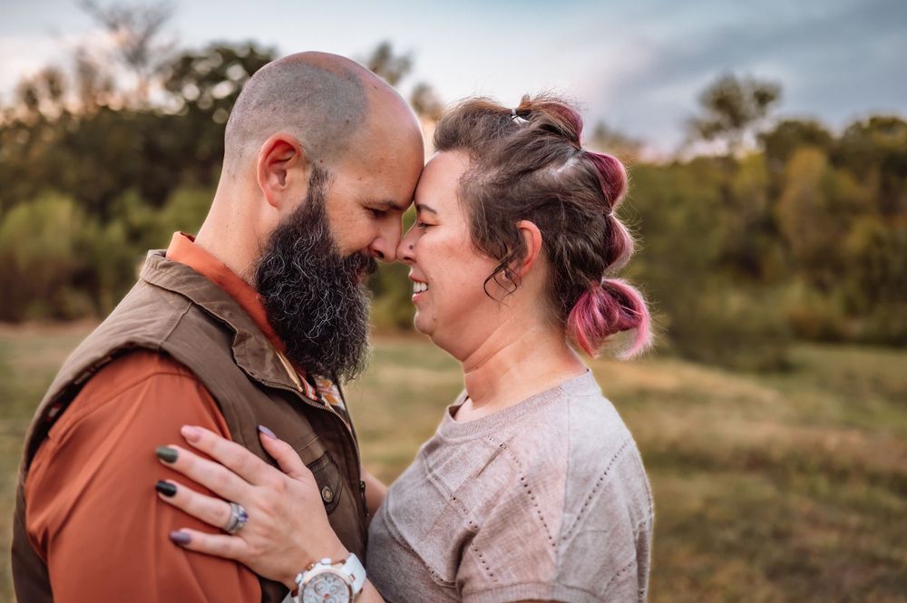 A bald bearded man and a woman with pink hair share a romantic forehead touch in a scenic outdoor field.