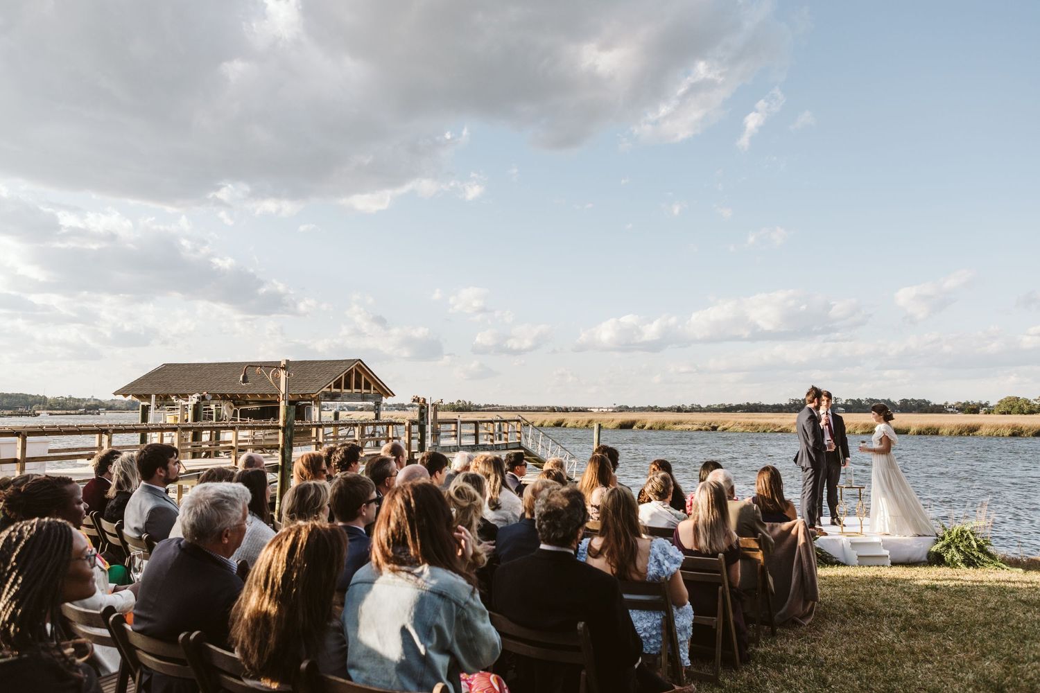 Outdoor waterfront wedding ceremony with guests gathered on sandy shore surrounded by tall pine trees under cloudy skies.