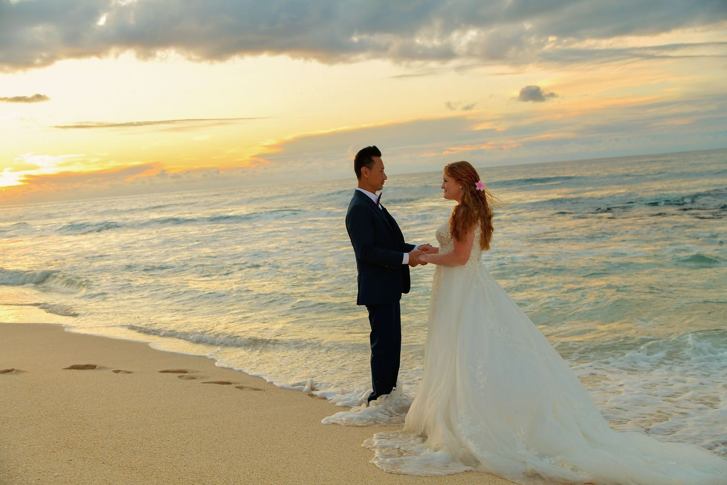 Couple exchanging vows on a sandy beach at sunset, bride in white gown and groom in navy suit by the ocean.