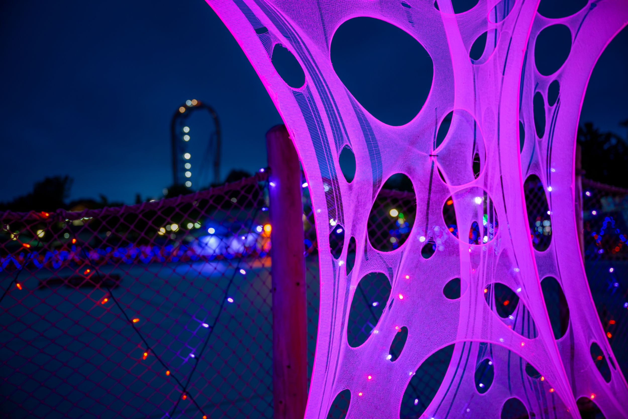 Glowing pink futuristic art installation with colorful lights at night, roller coaster visible in background.