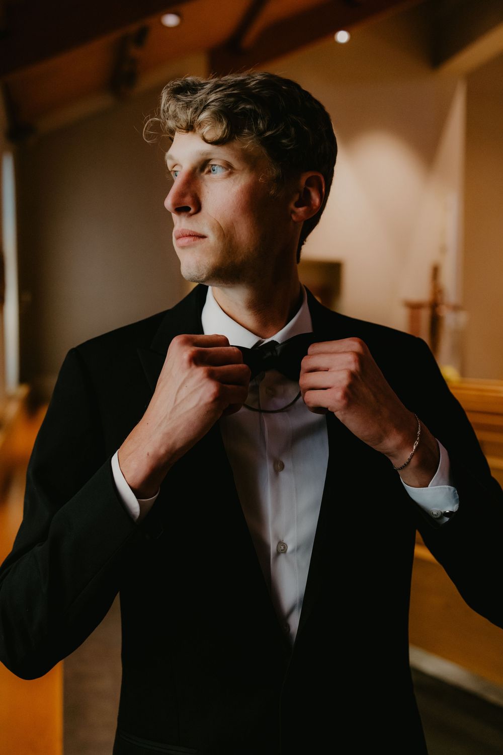 A young groom in a black tuxedo adjusts his bow tie while gazing away in an elegant indoor setting.