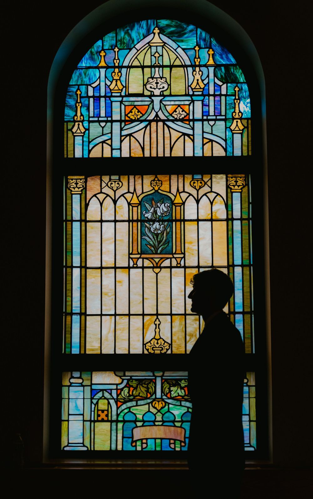 Silhouette of a person standing before a colorful stained glass church window with floral and architectural designs.