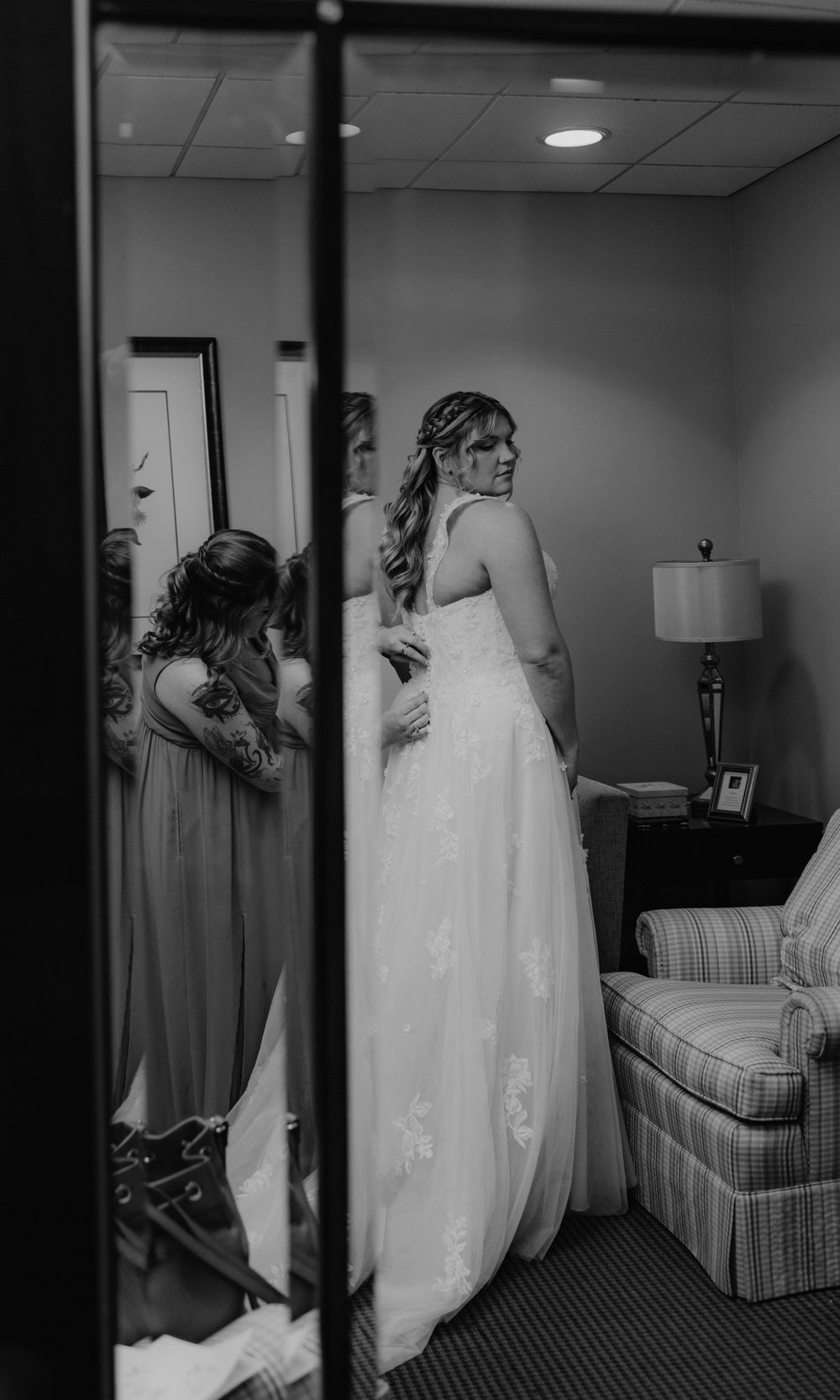 Black and white photo of a bride in a lace wedding gown being helped by a bridesmaid in a hotel room.