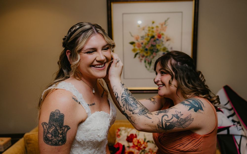 Two brides laughing joyfully together on wedding day, both showing colorful tattoos, in a warmly decorated room.