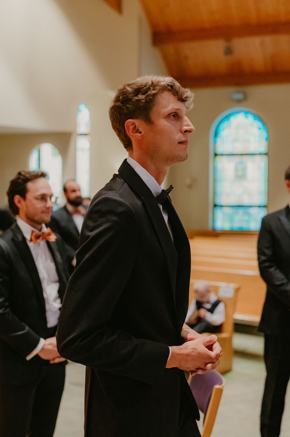 Groom in black tuxedo stands at church altar awaiting bride, with groomsmen in bow ties visible behind him.