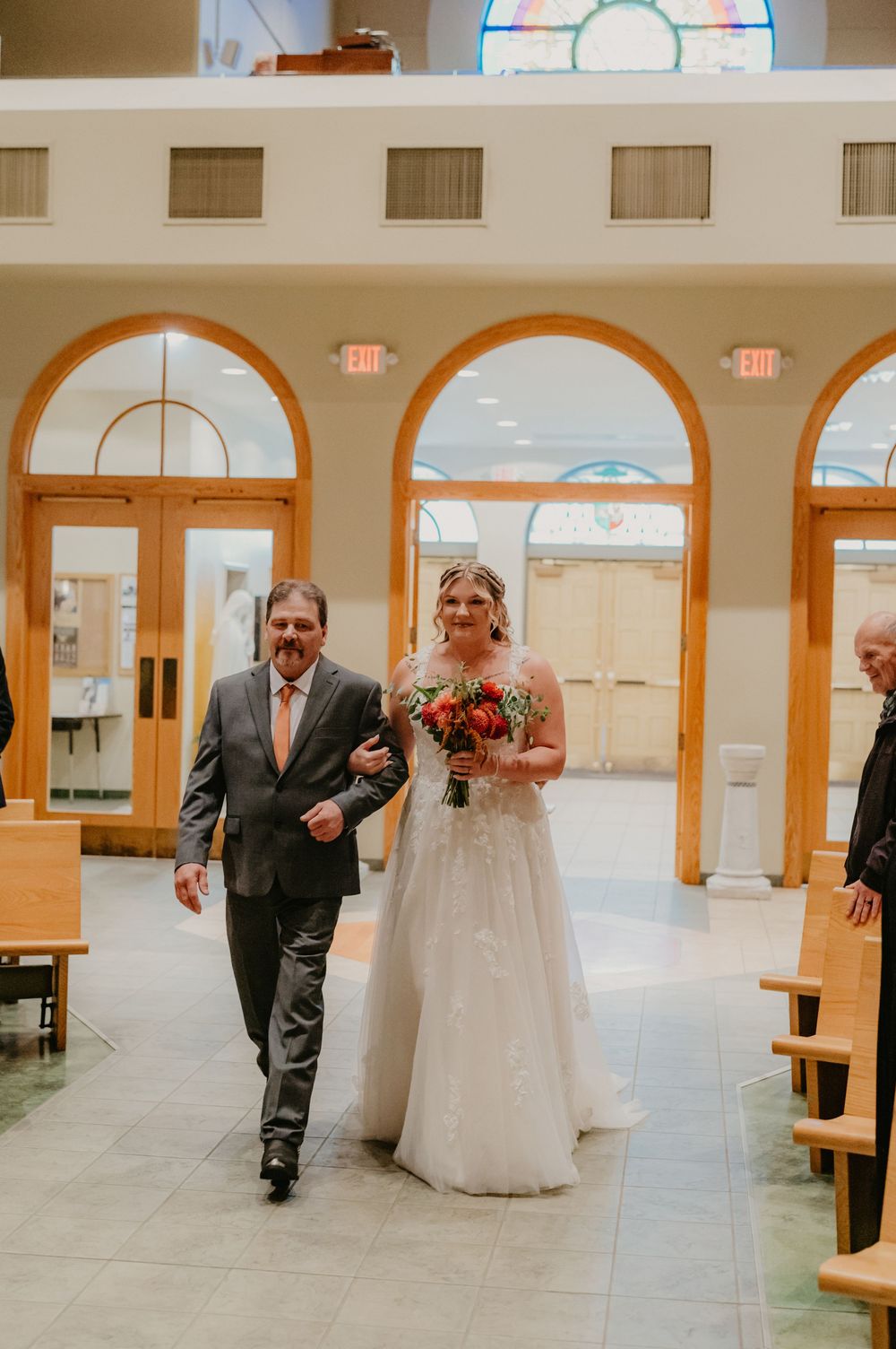 Bride in white gown walks down church aisle with escort, holding colorful bouquet, arched windows in background.
