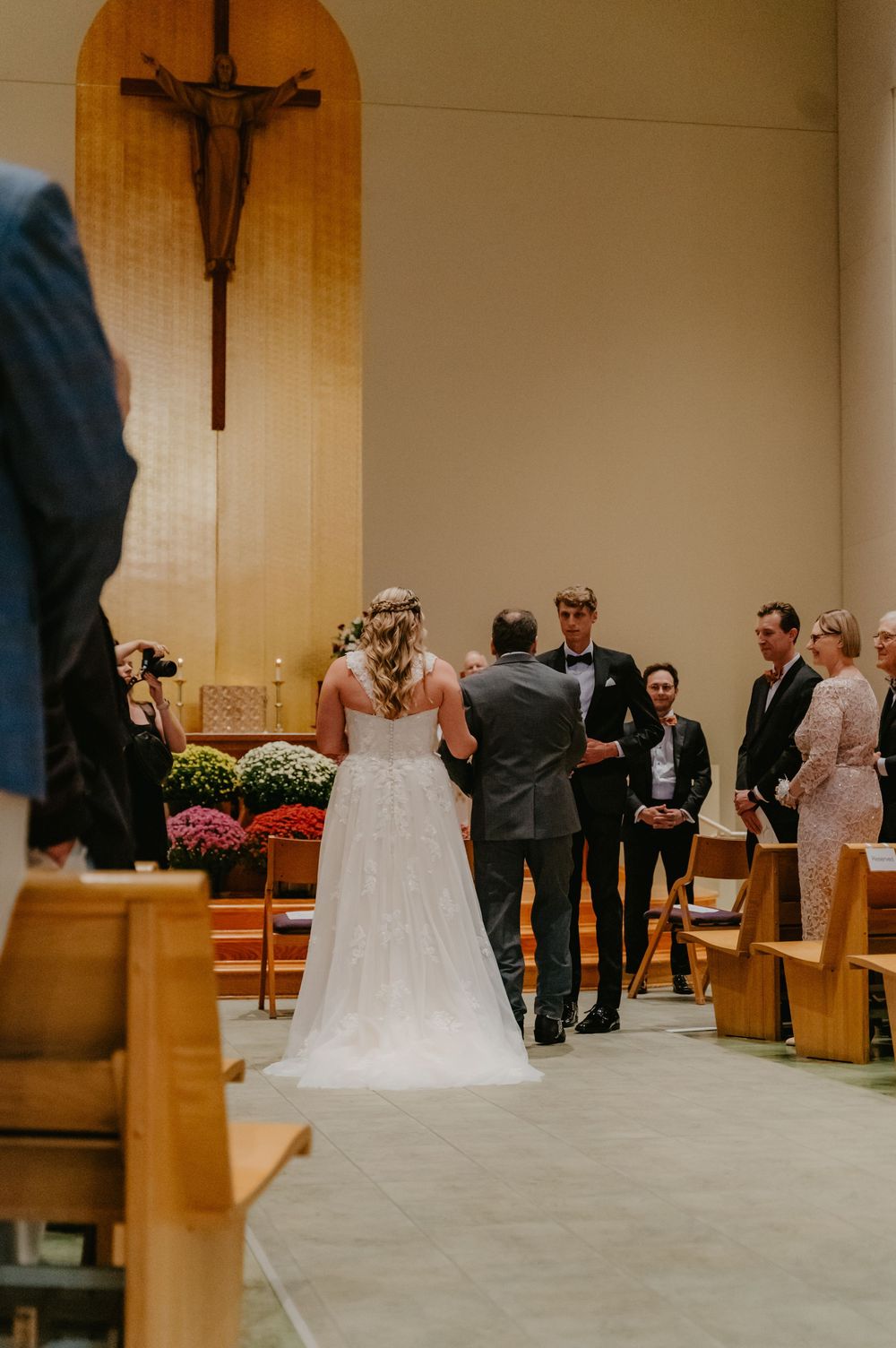 Bride in white gown walks down church aisle toward groom during wedding ceremony with cross altar backdrop.
