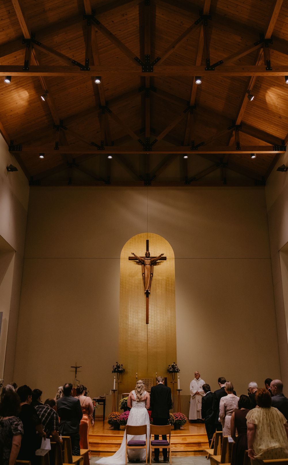 Wedding ceremony in a church with wooden ceiling, crucifix altar, bride and groom kneeling before priest.
