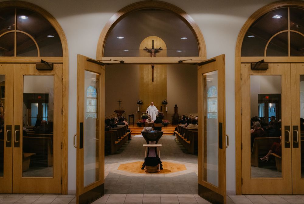 Interior view of a Catholic church sanctuary with wooden arched doors open to reveal altar, cross, and congregation during Mass.