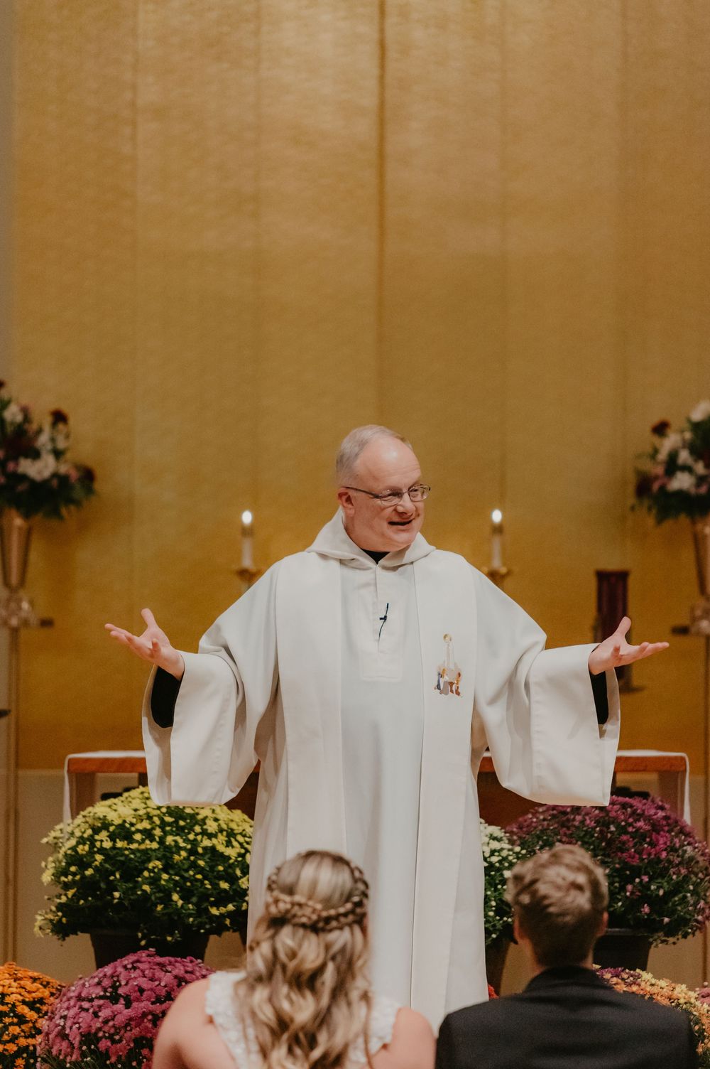 A priest in white robes officiates a wedding ceremony at a floral-decorated church altar with the couple seated before him.
