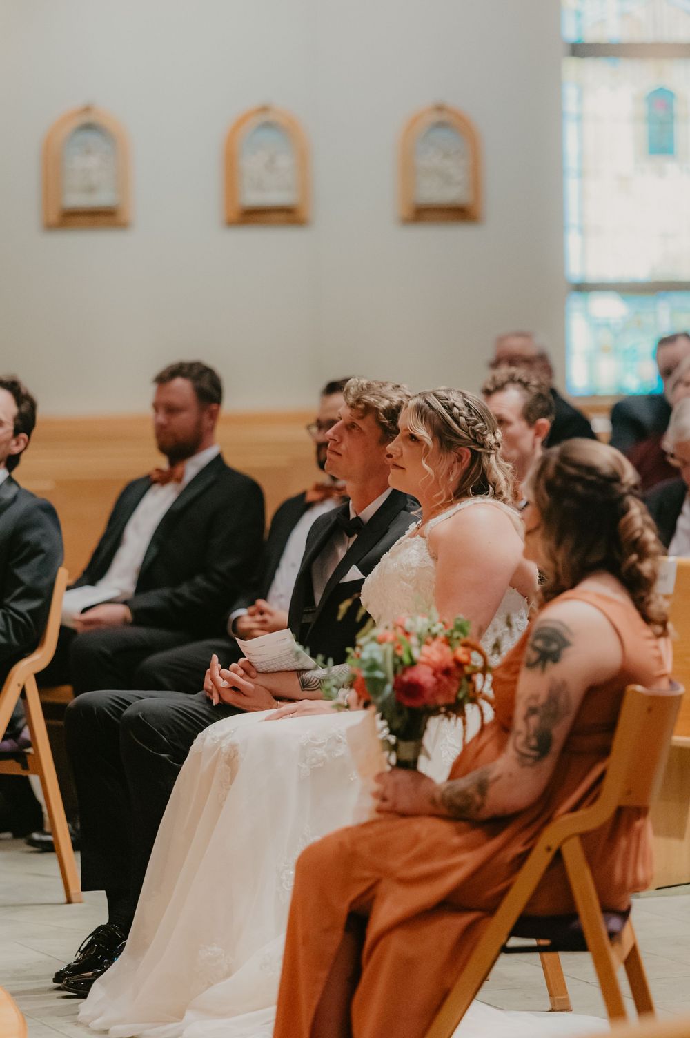 Bride and groom seated in church pews during wedding ceremony, surrounded by guests in formal attire.