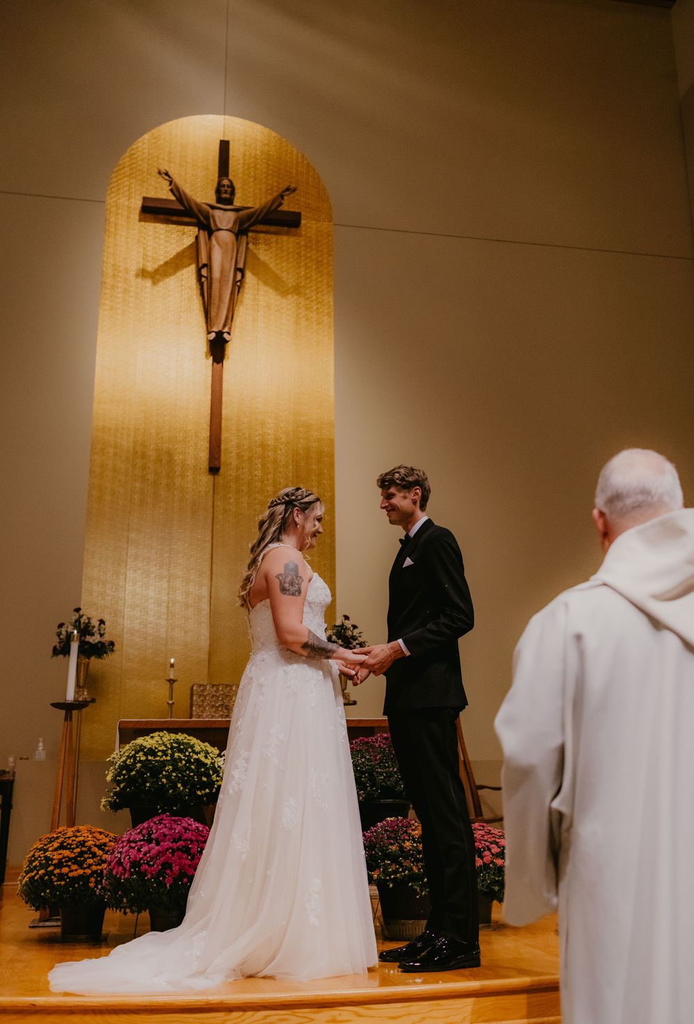 Bride and groom exchange vows at church altar with wooden crucifix, surrounded by colorful flowers, officiated by priest.