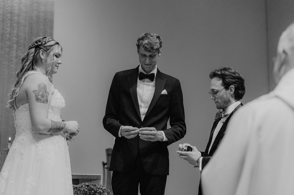 Black and white wedding ceremony photo of bride and groom exchanging rings with officiant.
