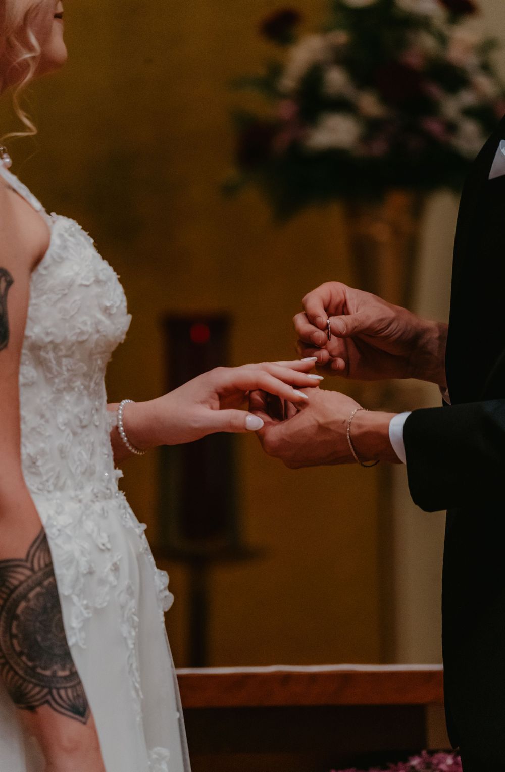 Groom placing wedding ring on bride's finger during ceremony, bride wearing white lace gown with floral tattoo visible.