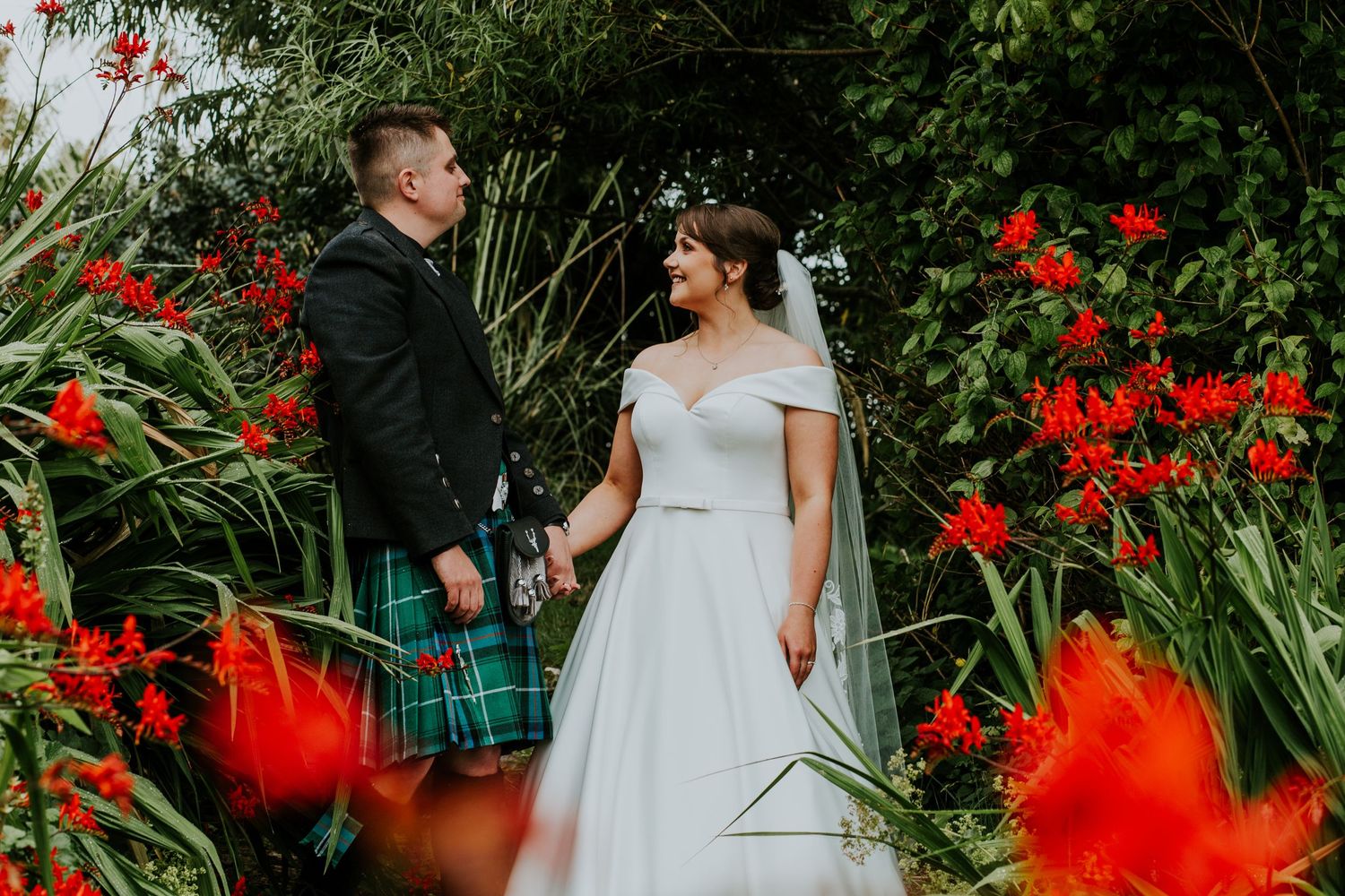 A romantic wedding couple poses in a garden with vibrant red flowers against lush greenery.