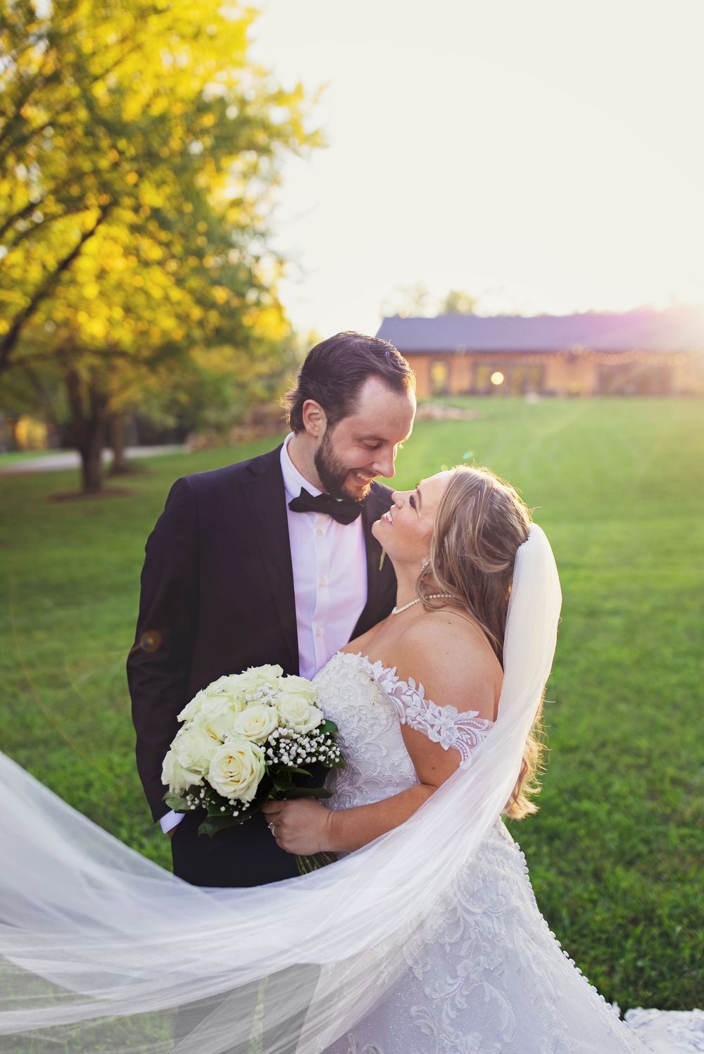 Backlit wedding portrait of a bride and groom smiling at each other. A bright golden hour sun flare illuminates a long veil.