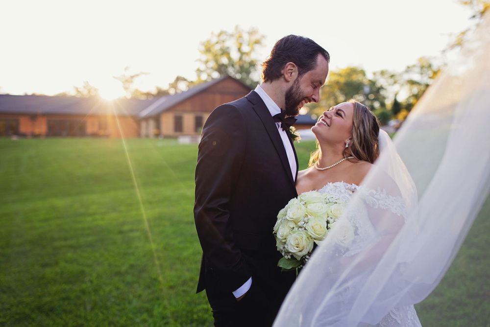 Backlit wedding portrait of a bride and groom smiling at each other. A bright golden hour sun flare illuminates a long veil.