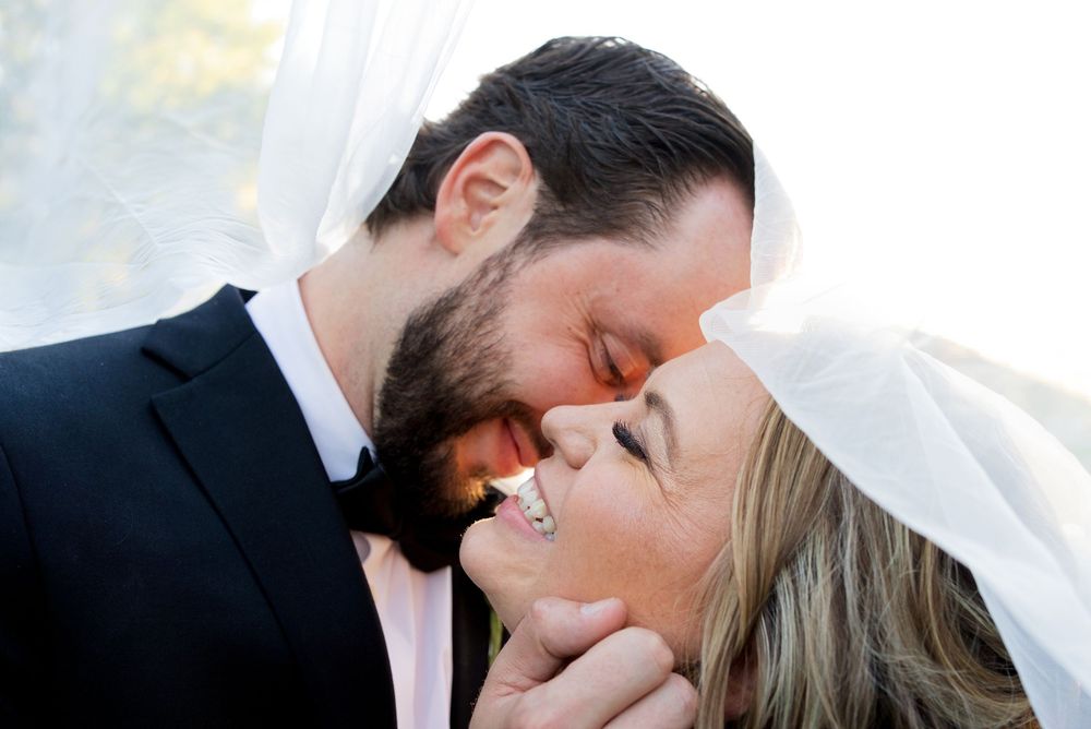 A close-up of the bride and groom under a veil. She is laughing while he caresses her face.